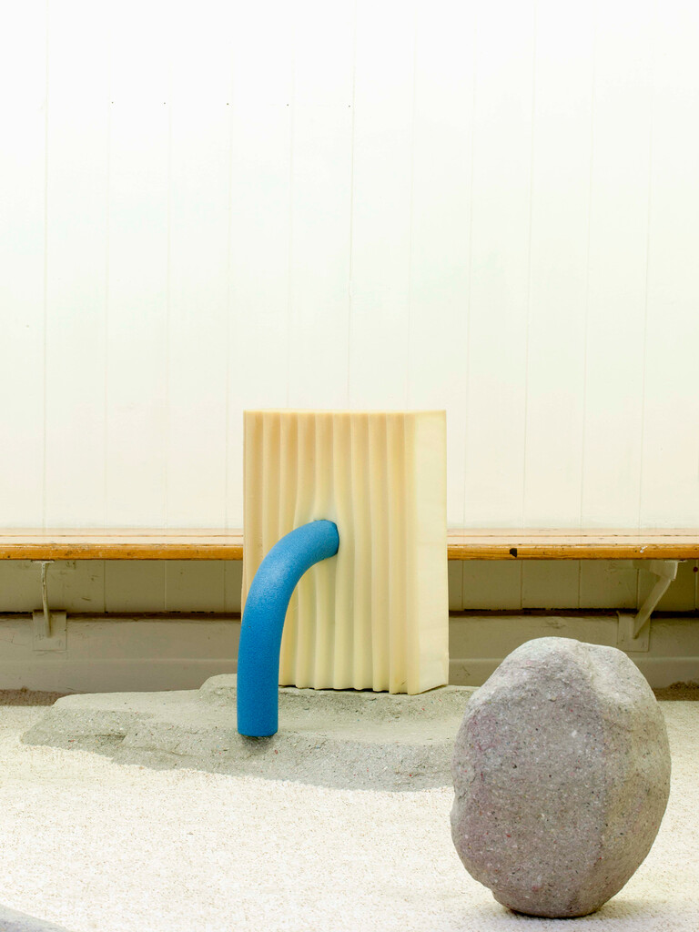 Ausstellungsansicht im Grand Palais Bern. Das Kunstwerk aus Schaumstoff ähnelt einem crèmefarbenen Ofen mit blauem Rohr. Es steht mit einem zweiten, steinähnlichen Kunstobjekt vor einer Sitzbank. Künstler: Jérôme Stünzi. Aus der Ausstellung «Une éternité provisoire», 2017. Foto: Guadalupe Ruiz.